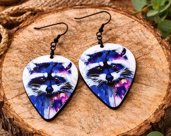 May include: A pair of teardrop-shaped earrings featuring a watercolor raccoon design. The earrings have black hooks and a white background with blue and purple accents. The earrings are displayed on a wooden surface.