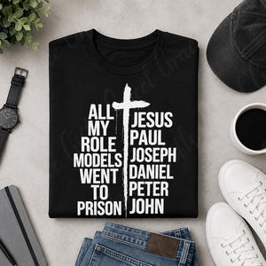May include: A black crew neck t-shirt featuring a white graphic of a cross separating two columns of text. The left side reads ALL MY ROLE MODELS WENT TO PRISON and the right side lists the names JESUS, PAUL, JOSEPH, DANIEL, PETER, and JOHN.