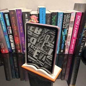May include: An e-reader with a pink border is displayed on a book-shaped stand, with a screen showing a jumbled letter design. A collection of books with colorful spines and titles like "King of Wrath" and "Bully" are in the background. A small pink figurine sits atop the books.