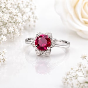 Op de afbeelding: Een zilveren ring met een bloemvormige zetting. De centrale steen is een grote, ronde, dieprode edelsteen, omringd door kleinere, heldere accentstenen. De ring staat tegen een witte achtergrond met witte bloemen.