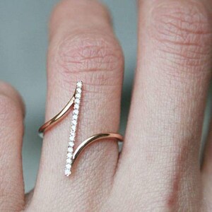 Könnte beinhalten: Ein roségoldener Ring mit einer vertikalen Reihe kleiner, klarer Edelsteine. Der Ring hat ein einzigartiges Design mit einem geschwungenen Band, das den Finger umschließt und mit dem mit Edelsteinen besetzten Stab verbunden ist.