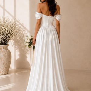 May include: A floor-length white satin wedding dress featuring an off-the-shoulder neckline with draped sleeves, a fitted bodice with a vertical row of small fabric-covered buttons down the centre back, and a full, flowing skirt with a train.