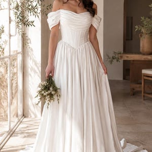 May include: A floor-length white gown featuring an off-the-shoulder neckline with draped fabric, a structured corset-style bodice with vertical boning, and a full, pleated skirt that extends into a train, held with a small bouquet of white flowers.