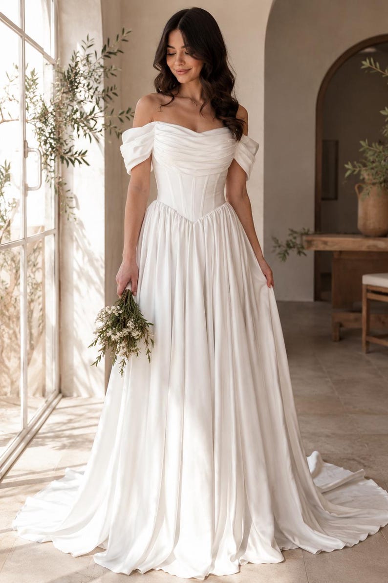 May include: A floor-length white gown featuring an off-the-shoulder neckline with draped fabric, a structured corset-style bodice with vertical boning, and a full, pleated skirt that extends into a train, held with a small bouquet of white flowers.