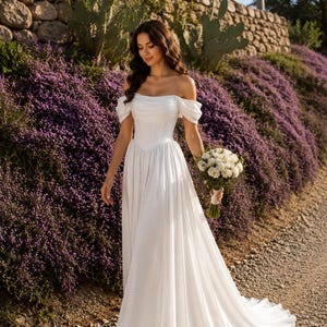 May include: A floor-length white wedding dress featuring an off-the-shoulder neckline with draped sleeves, a fitted bodice with a pointed waistline, and a flowing skirt. The dress is paired with a bouquet of white flowers and silver high-heeled sandals.