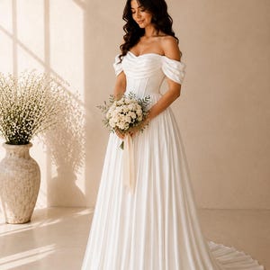May include: A floor-length white wedding dress featuring an off-the-shoulder neckline with draped fabric, a structured corset-style bodice, and a full, pleated skirt that extends into a train. The dress is made of a smooth, satin-like material.