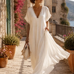 May include: A flowing white dress with a V-neck and lace trim. The dress is worn with sandals and a patterned handbag. The scene is set on a cobblestone street with flowering plants and stone buildings, creating a relaxed, bohemian aesthetic.