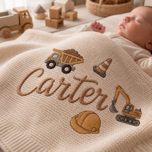 May include: A cream-coloured knit baby blanket featuring embroidered construction-themed designs including a dump truck, an excavator, a traffic cone, and a hard hat. The name Carter is embroidered in brown script across the centre of the blanket.