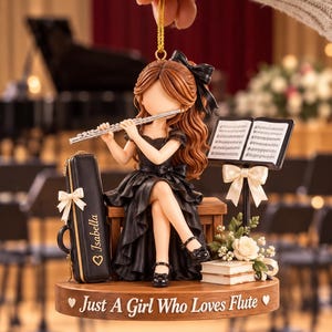 May include: A hanging ornament featuring a brown-haired figure in a black dress playing a silver flute while seated on a bench. Beside the figure is a black case labelled Isabella, a music stand with sheet music, and a base inscribed Just A Girl Who Loves Flute.