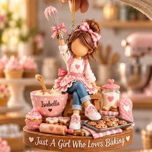 May include: A decorative ornament featuring a figurine of a person with brown hair and a pink gingham bow, wearing a white shirt and pink apron, sitting on a stool. The base is inscribed with "Just A Girl Who Loves Baking" and includes a bowl labeled "Isabella".
