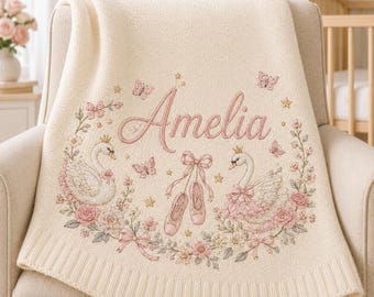 Peut inclure: Une couverture en tricot crème avec le prénom Amelia brodé en écriture rose. Le motif comprend deux cygnes portant des couronnes, des chaussons de danse roses avec un nœud, des papillons, des étoiles et une couronne de roses et feuilles.