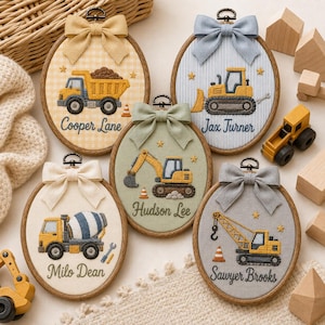 May include: Five oval wooden embroidery hoops featuring fabric backgrounds in yellow gingham, blue stripes, cream, sage green, and gray. Each hoop displays a stitched construction vehicle, a fabric bow at the top, and a personalized name in cursive script.