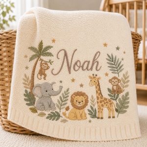 May include: A cream-coloured knit blanket featuring an embroidered jungle scene with a palm tree, a monkey, a grey elephant, a lion, and a giraffe. The name Noah is embroidered in brown script in the centre, surrounded by small stars and leaves.