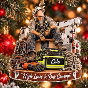 May include: A clear acrylic ornament featuring a lineman sitting on a wooden spool, wearing a hard hat, safety harness, and work gloves. A tool bag labeled "Cole" sits in front, with the text "High Lines & Big Courage" printed below.