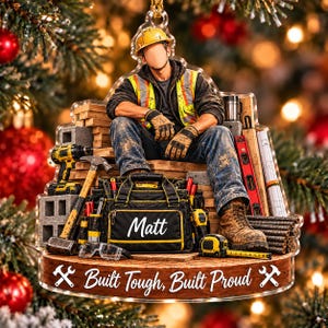 May include: A clear acrylic ornament featuring a construction worker in a yellow hard hat, high-visibility vest, and work boots. A tool bag labeled Matt sits in front, surrounded by tools, wood, and the text Built Tough, Built Proud.