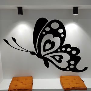 Peut inclure: Un grand autocollant de papillon noir est fixé sur un mur blanc, avec un motif d'ailes détaillé. Deux coussins orange sont placés en dessous. Deux spots noirs sont montés au plafond.
