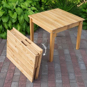 Puede incluir: Una mesa cuadrada de madera clara con una mesa plegable a juego. La mesa tiene una tapa de listones y cuatro patas. La mesa plegable está parcialmente plegada, mostrando los listones de madera y las asas recortadas. Las mesas están al aire libre.