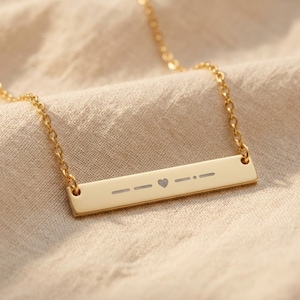 May include: A gold-plated bar necklace with a fine chain. The rectangular pendant has engraved Morse code symbols and a heart symbol. The necklace is shown on a neutral-toned fabric.