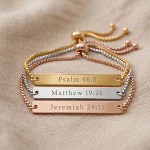 Puede incluir: Tres pulseras ajustables en oro, plata y oro rosa. Cada pulsera presenta una barra rectangular grabada con un versículo bíblico: Salmo 46:5, Mateo 19:26 y Jeremías 29:11. Las pulseras se exhiben sobre una tela de tono neutro.