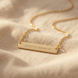 May include: A gold-colored bar necklace with a delicate chain. The rectangular pendant is engraved with the coordinates 25.7617° N • 80.1918° W. The necklace rests on a beige fabric surface.