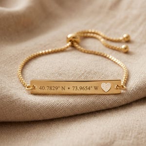 May include: Gold-coloured bracelet featuring a rectangular bar engraved with coordinates and a heart-shaped cutout. The adjustable chain bracelet is shown on neutral fabric. The coordinates are 40.7829° N • 73.9654° W.