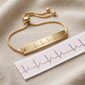 Puede incluir: Una pulsera dorada con una barra rectangular grabada con un patrón de electrocardiograma. La pulsera está sobre una superficie beige junto a una tira de papel con un gráfico de ECG similar. La pulsera tiene un cierre deslizante.