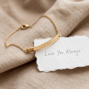 May include: A gold-colored bracelet with a curved bar pendant. The pendant is inscribed with "Love You Always." The bracelet is on a neutral-toned fabric, next to a handwritten note that says "Love You Always."