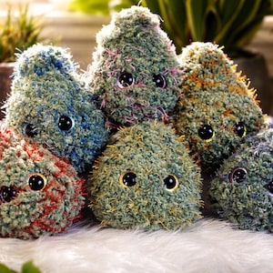 May include: Six handmade plush toys with fuzzy, textured bodies in shades of green, blue, and red. Each has large, black, googly eyes. The toys have a whimsical appearance. They are arranged on a soft, white surface, creating a playful scene.