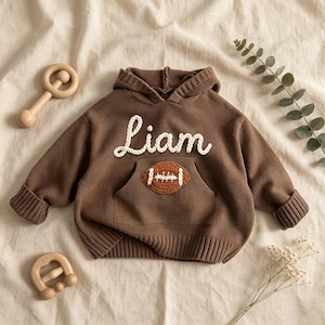 Könnte beinhalten: Ein brauner, langärmeliger Strick-Kapuzenpullover mit dem Namen Liam in weißer Schreibschrift auf der Brust und einer gehäkelten Football-Applikation auf der Kängurutasche, ausgestattet mit gerippten Bündchen an Ärmeln und Saum.