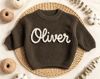 Könnte beinhalten: Ein dunkelgrüner Strickpullover mit dem Namen "Oliver" in weißer Schrift. Der Pullover wird auf einer gewebten Matte präsentiert, umgeben von Wollknäueln und Baumwollblüten. Der Pullover hat lange Ärmel und einen Rundhalsausschnitt.