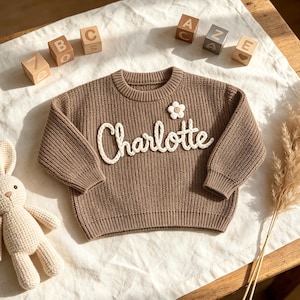 May include: A brown knitted sweater with the name "Charlotte" in white script. A small white flower is attached near the neckline. Wooden alphabet blocks and a stuffed bunny are in the background.