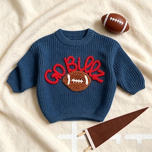 May include: A dark blue long-sleeve knit jumper featuring the red embroidered text GO BILLY and a brown football applique with white stitching. A separate brown football and a brown felt pennant are positioned near the jumper.
