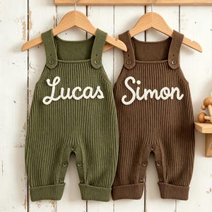 Könnte beinhalten: Zwei Baby-Latzhosen, eine olivgrüne und eine braune, hängen an Holzbügeln. Die olivgrüne Latzhose trägt den Namen "Lucas" in weißer Schrift, während die braune "Simon" trägt. Beide sind gerippt gestrickt und haben Knöpfe.