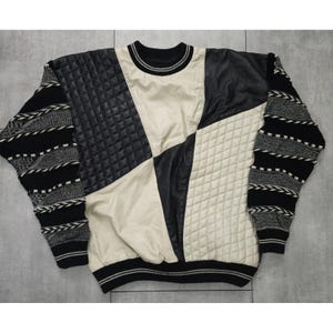 Può includere: Un maglione a maniche lunghe con un design a blocchi di colore caratterizzato da pannelli neri trapuntati, pannelli lisci color crema e maniche in maglia testurizzata nera e grigia. Il capo presenta un girocollo e polsini e orlo a coste.