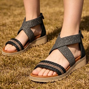 May include: Sandals with a black and gray crisscross strap design. The sandals have a black braided strap across the toes with gold-colored accents. The soles are tan and the background is brown grass.