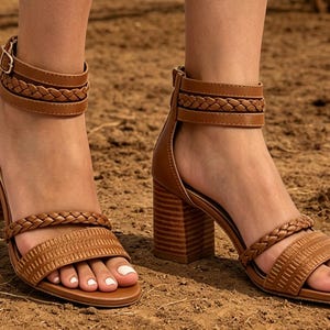 Könnte beinhalten: Braune Ledersandalen mit Blockabsatz und geflochtenen Riemen. Die Sandalen haben einen Knöchelriemen mit Schnallenverschluss und einen Reißverschluss hinten. Das offene Design zeigt weiß lackierte Zehennägel.