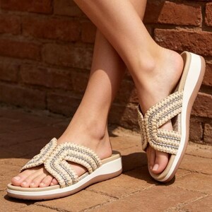 May include: Beige and tan woven sandals with a crisscross strap design. The sandals have a white sole and a brown bottom sole. The sandals are on a brick surface.