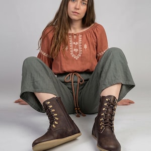May include: A pair of dark brown leather boots with a side zipper and gold-colored eyelets. The boots have a flat sole and are laced up the front. The boots are part of a barefoot shoe collection.