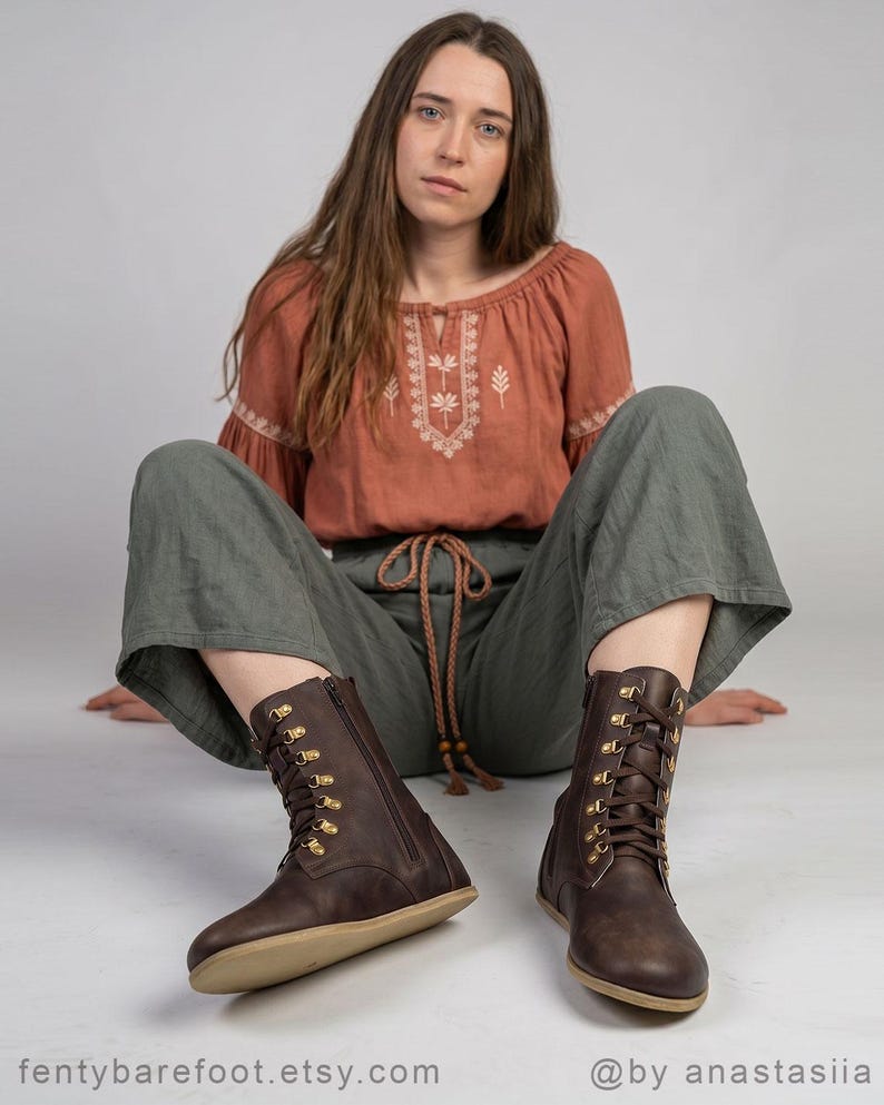 May include: A pair of dark brown leather boots with a side zipper and gold-colored eyelets. The boots have a flat sole and are laced up the front. The boots are part of a barefoot shoe collection.