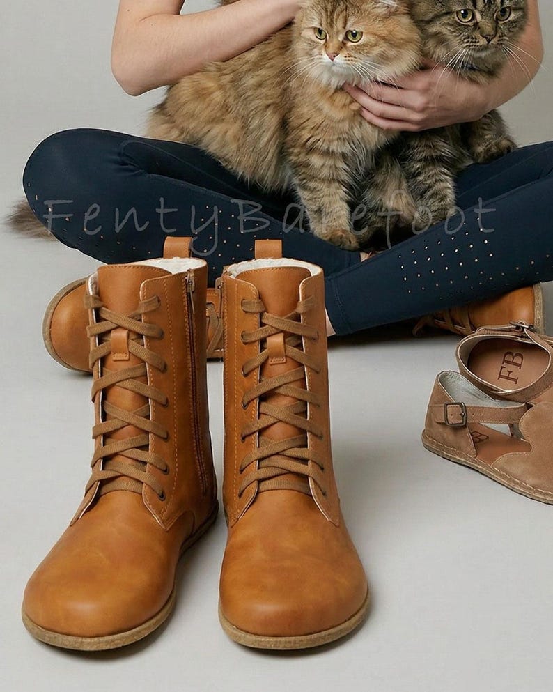 May include: A pair of tan leather lace-up boots with side zippers. The boots have a rounded toe and a flat sole. A pair of brown sandals with a buckle are also visible. The image also shows a person holding two cats.