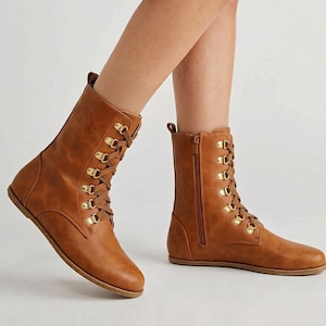 May include: A pair of brown leather boots with a flat sole. The boots have a lace-up design with gold-colored eyelets and a side zipper. The boots are mid-calf height.