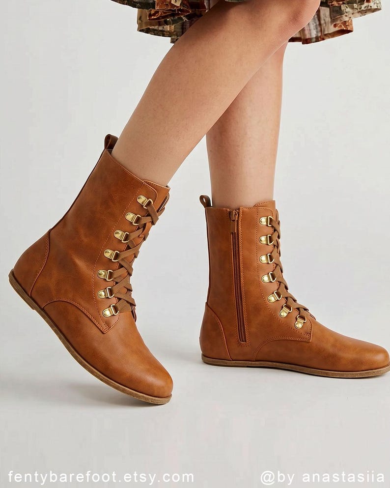 May include: A pair of brown leather boots with a flat sole. The boots have a lace-up design with gold-colored eyelets and a side zipper. The boots are mid-calf height.