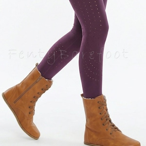 Op de afbeelding: Bruine leren laarzen met veters en een zijrits, gecombineerd met donkerpaarse leggings met kleine stippeluitsparingen. De laarzen hebben een platte zool. De tekst "Fenty Barefoot" is zichtbaar.
