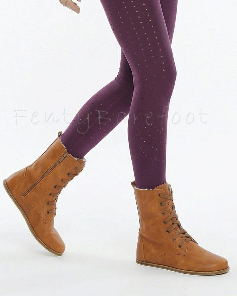 Op de afbeelding: Bruine leren laarzen met veters en een zijrits, gecombineerd met donkerpaarse leggings met kleine stippeluitsparingen. De laarzen hebben een platte zool. De tekst "Fenty Barefoot" is zichtbaar.