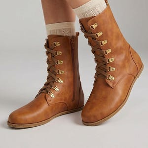 May include: Brown leather boots with a lace-up design and gold-tone eyelets. The boots have a side zipper and are worn with cream-colored socks. The boots are flat-soled and have a classic, rugged style. The Etsy shop name is visible at the bottom of the image.