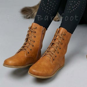Op de afbeelding: Bruin gekleurde veterlaarzen met zijritsen. De laarzen zijn gemaakt van een glad materiaal en hebben een platte zool. De laarzen worden gedragen met zwarte leggings met gouden stippen. De tekst "Fenty Barefoot" is zichtbaar.