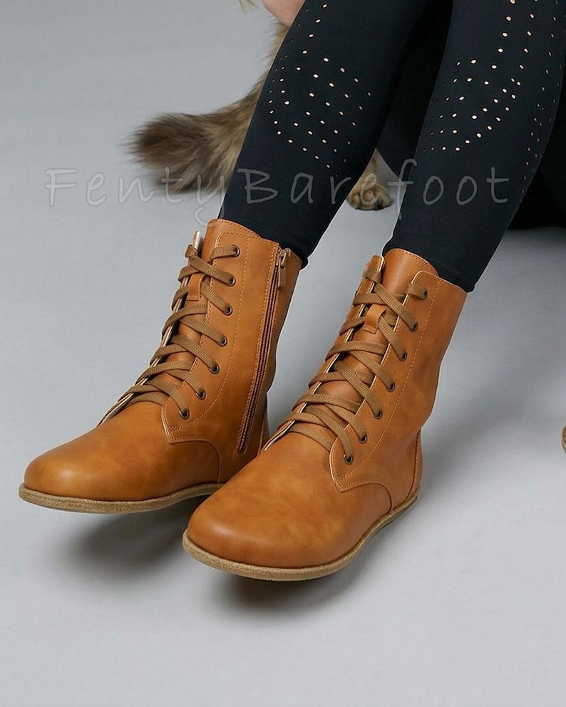 Op de afbeelding: Bruin gekleurde veterlaarzen met zijritsen. De laarzen zijn gemaakt van een glad materiaal en hebben een platte zool. De laarzen worden gedragen met zwarte leggings met gouden stippen. De tekst "Fenty Barefoot" is zichtbaar.