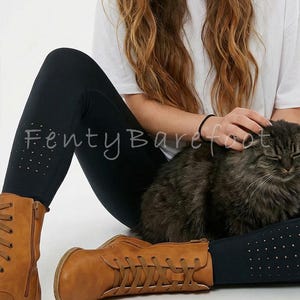 Op de afbeelding: Zwarte legging met geperforeerde details en bruine leren laarzen met veters en ritsen. De tekst "Fenty Barefoot" is zichtbaar op de legging. Een pluizige kat ligt op schoot.