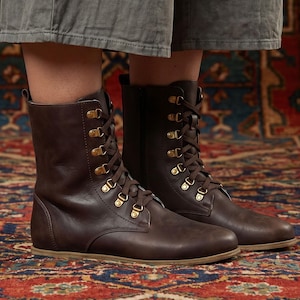 May include: Brown leather boots with brass eyelets and brown laces. The boots have a flat sole and a mid-calf height. The boots are displayed on a patterned rug. The Etsy shop name "fentybarefoot.etsy.com" is visible at the bottom of the image.