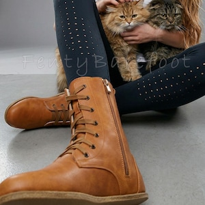 Op de afbeelding: Lichtbruine veterlaarzen met een zijrits. De laarzen worden gedragen met donkerblauwe leggings met kleine cirkelvormige uitsparingen. Twee katten worden op schoot gehouden. De tekst "FentyBarefoot" is zichtbaar.